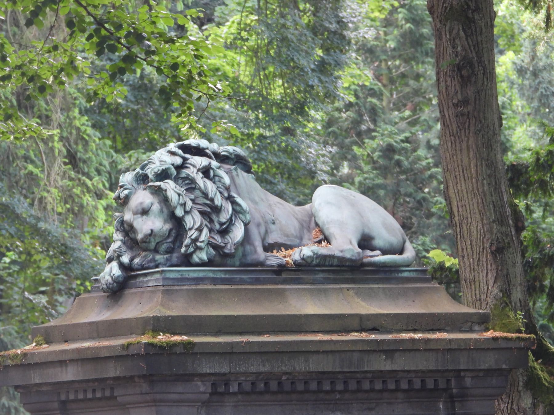 Ruhende bronzene Löwenfigur auf hohem Steinsockel zwischen alten Bäumen auf Hauptfriedhof Mainz